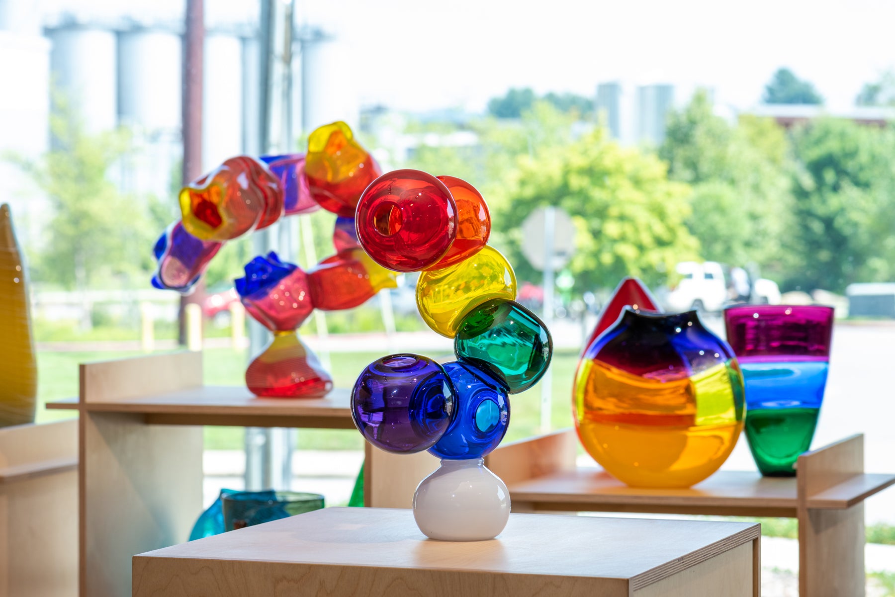A view of a rainbow colored bubble sculpture in the small batch glass gallery with more brightly colored glass vases in the background. Image by Loam