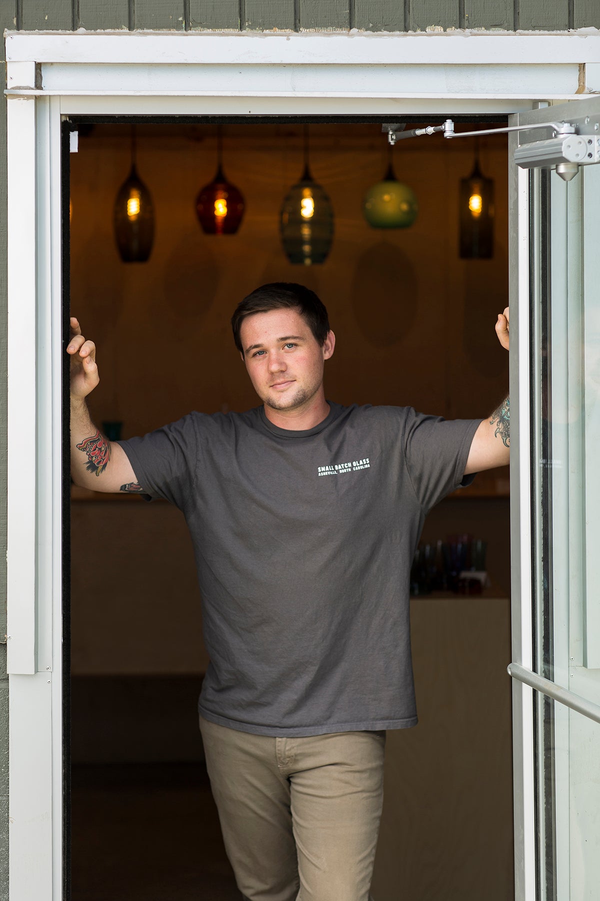 Artist and owner of Small Batch Glass Blowing stands in the doorway of his gallery wearing a grey shirt and khaki pants, arms propped on the doorframe. 