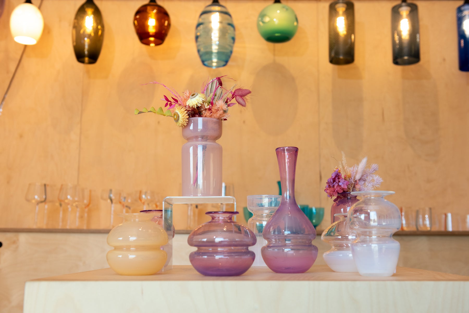 A view inside the gallery of Small Batch Glass where several pink shade bud vases sit on a pedestal in front of hanging lights. Image by Loam