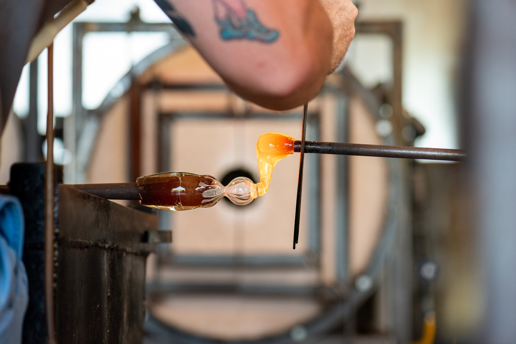 A close up view of molten glass being added to a nub of glass with a blurry Small Batch Glass hot shop background. Image by Loam