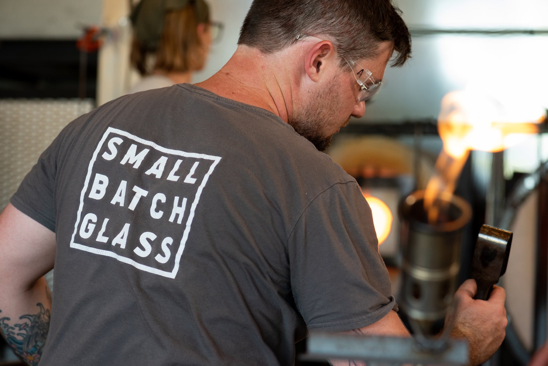 Asher Holman, the main glassblower at Small Batch Glass, from the back with flames in the foreground and a glowing furnace behind him. Image by Loam