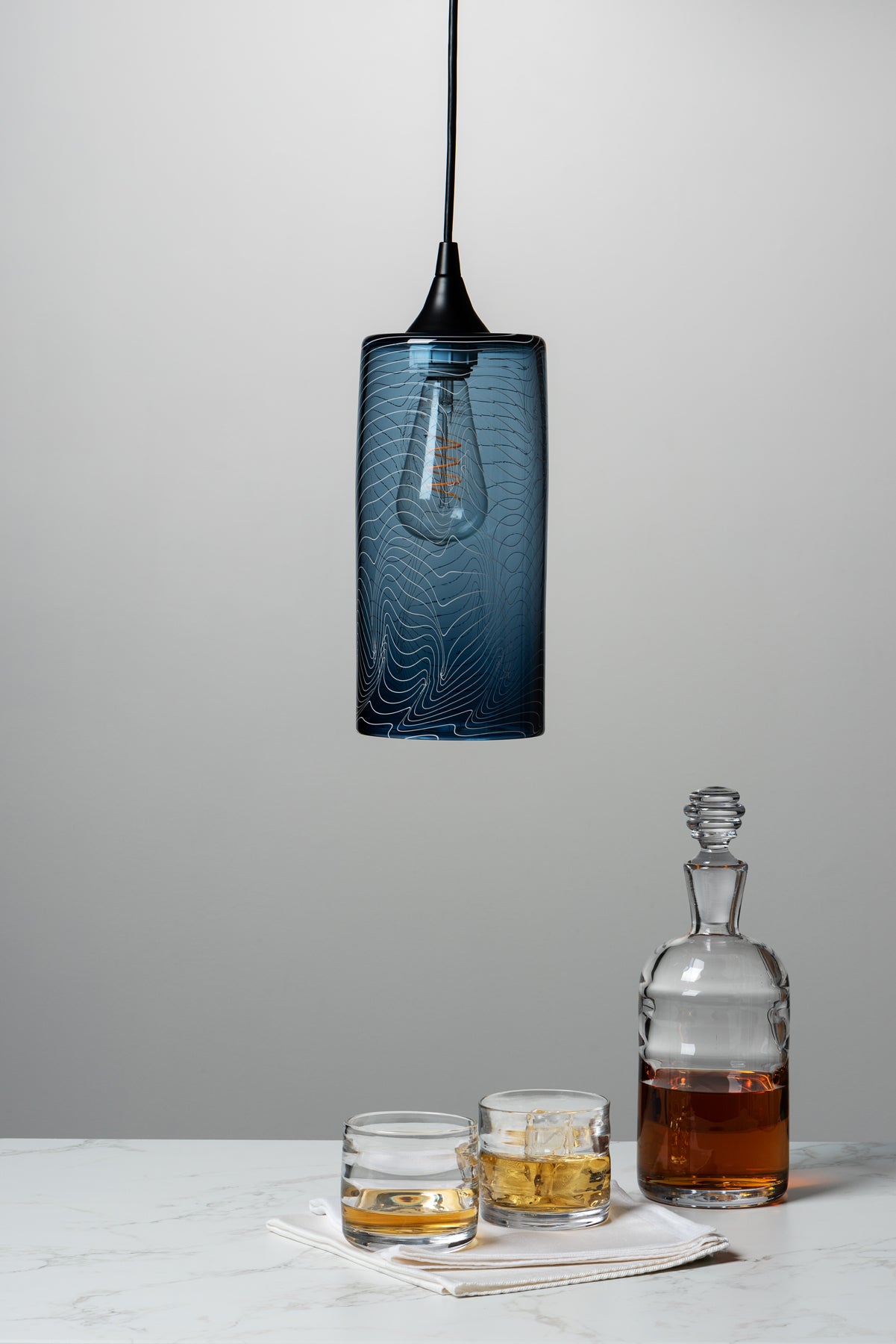 A small batch glass light fixture hangs above a marble table top where a decanter and rocks glasses sit against a grey background. Image by Loam