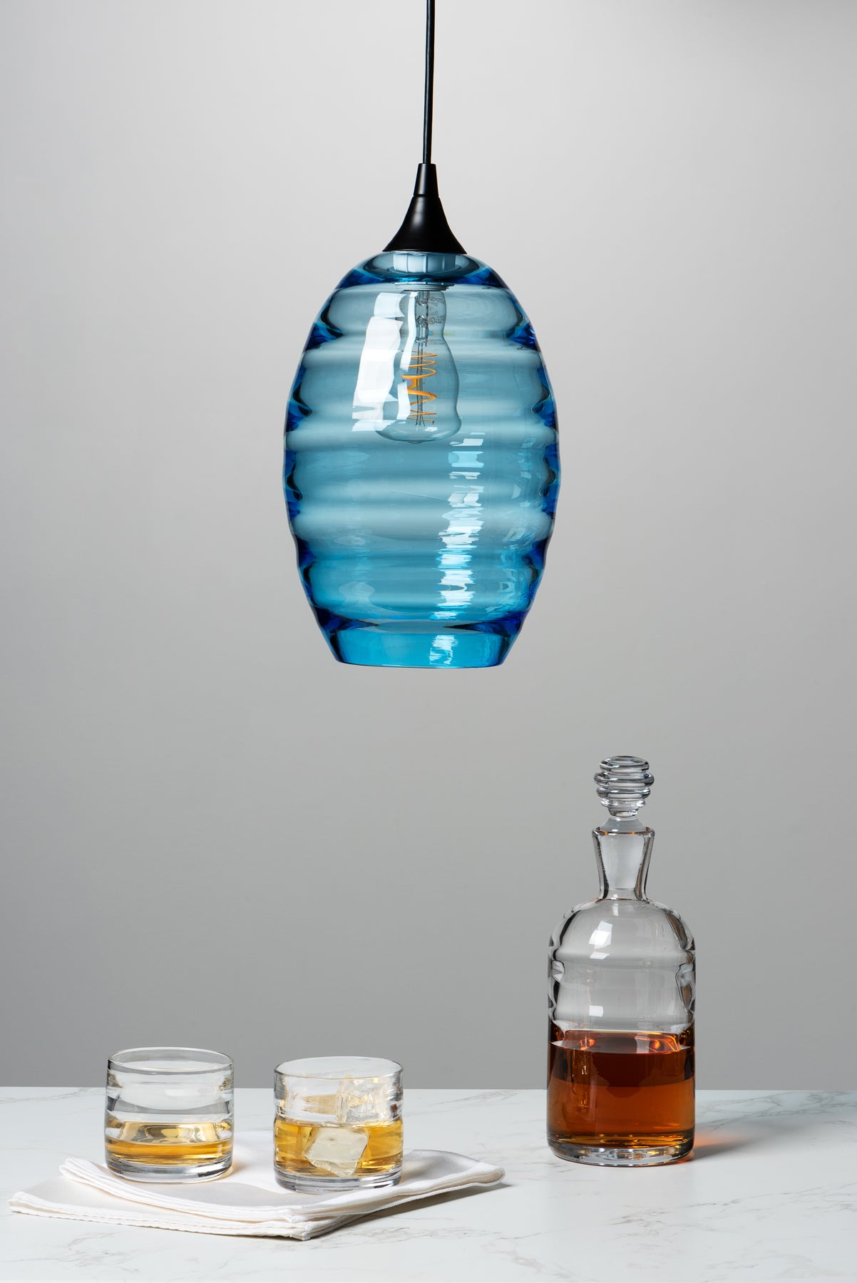 The Small Batch Glass Lighthouse series decanter, rocks glasses, and pendant light are in the frame - the decanter and glasses sit on the table and the light hands from above. Image by Loam