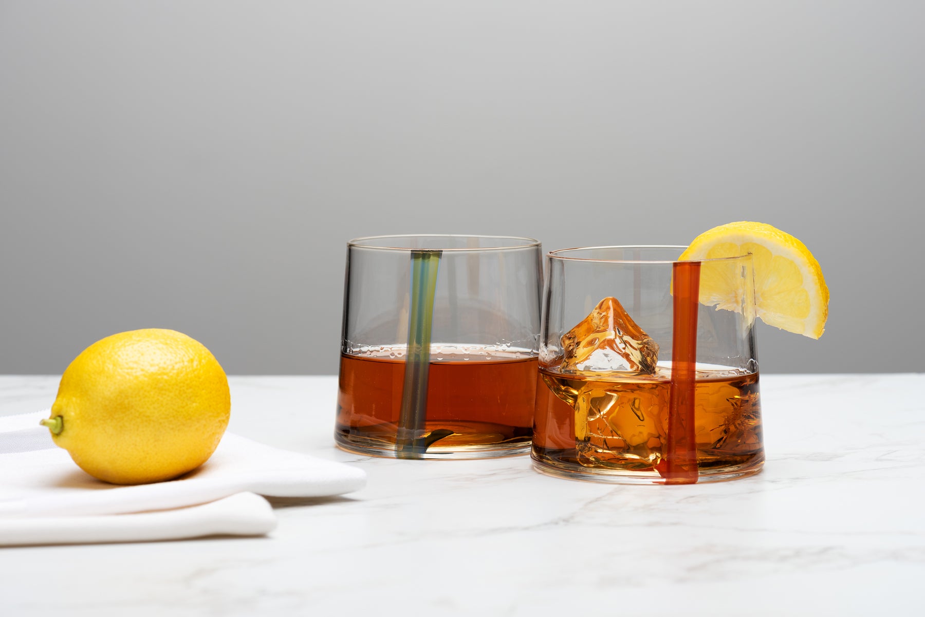 Two straight sided rocks glasses with a stripe of color on each, sit atop a marble table with a lemon. Image by Loam