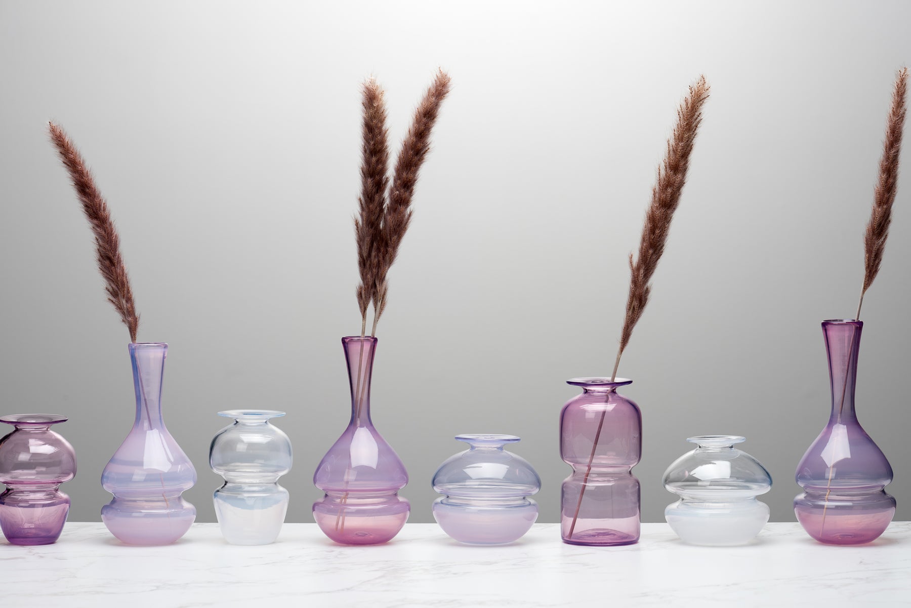 A line up of pink and purple shaded Small Batch Glass bud vases in many different shapes. Image by Loam