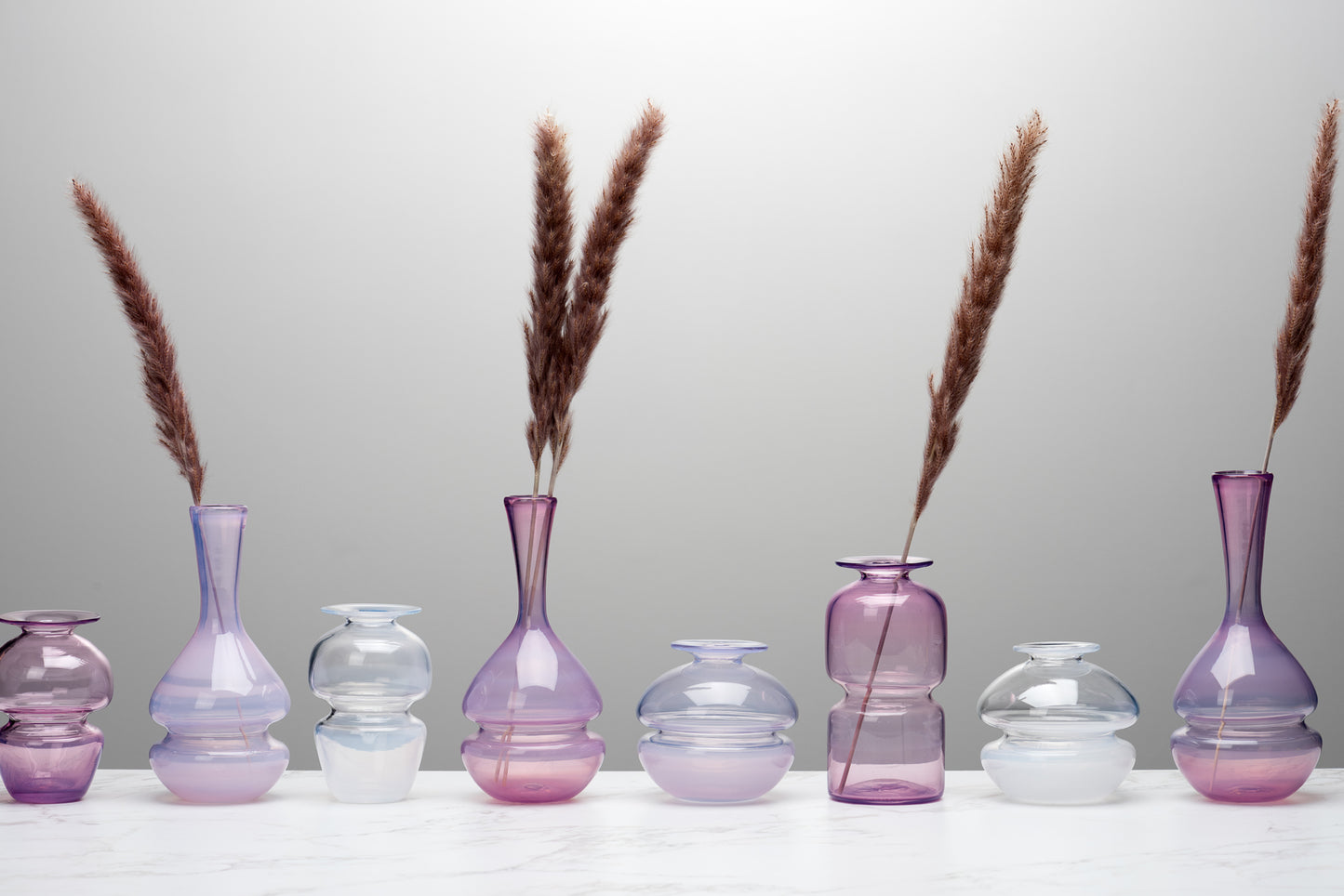 A line-up of pink purple and white bud vases holding grasses. They sit atop a marble table against a grey background. Image by Loam